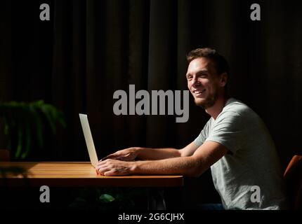 Side view of smiling young man sitting in cafe and working or studying in laptop. Concept of process working at home or cafe with good mood. Stock Photo