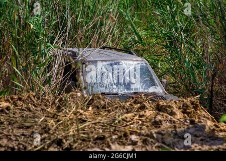on the river bank a sunken SUV with a dirty windshield in the reeds, a rally racing car is mired in a lake swamp, nobody. Stock Photo