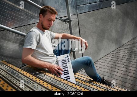 Side view of young man sitting on staircase and playing small white digital piano. Concept of time on street with good mood and sunny weather. Stock Photo