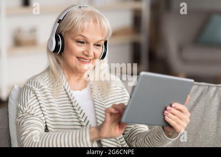 Smiling mature woman in headset using tablet sitting on sofa Stock Photo