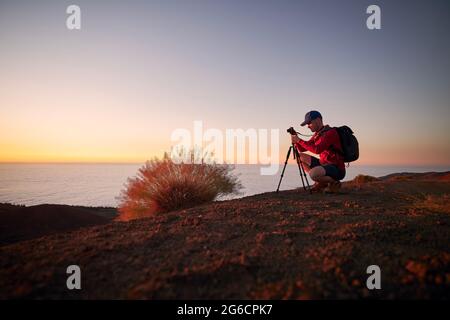 Young man during photographing landscape on top of hill above clouds. Hobby photographer waiting for beautiful sunset. Tenerife, Canary Islands, Spain Stock Photo