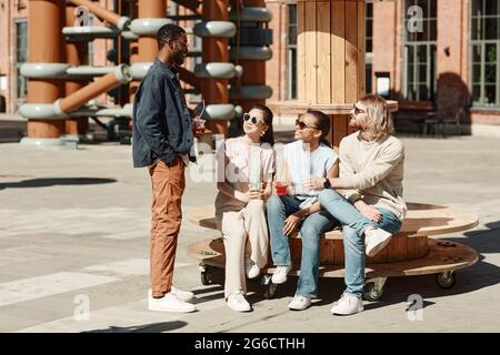 Full length portrait of diverse group of young people chatting while rrelxing in urban city setting Stock Photo