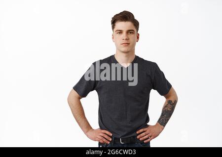 Confident young man standing with hands on waist, smiling and looking determined, ready for work, waiting for task, posing in t-shirt and jeans, white Stock Photo