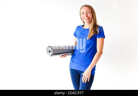 Blond slim female in bright blue top and leggings unrolling mat for yoga practice in white studio Stock Photo