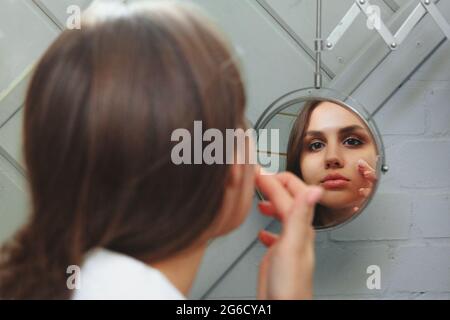 Young female in white bathrobe smearing mud mask on face while standing in front of mirror during spa procedure at home Stock Photo