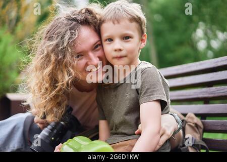 Delighted loving Woman embracing cute boy while sitting on bench in park and looking at camera Stock Photo