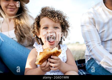 Cheerful little girl eating pastry looking at camera sitting with multiracial family enjoying picnic together in nature Stock Photo
