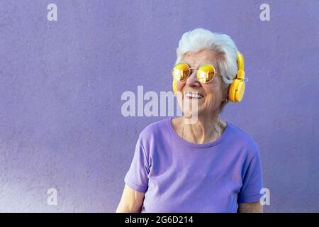 Cheerful senior female in t shirt and modern sunglasses listening to song from wireless headset on purple background Stock Photo