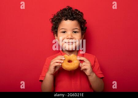 Adorable child with curly hair eating sweet tasty doughnut and looking at camera on red background Stock Photo