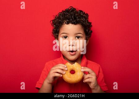 Adorable child with curly hair eating sweet tasty doughnut and looking at camera on red background Stock Photo