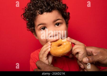 Adorable child with curly hair eating sweet tasty doughnut and looking at camera on red background Stock Photo