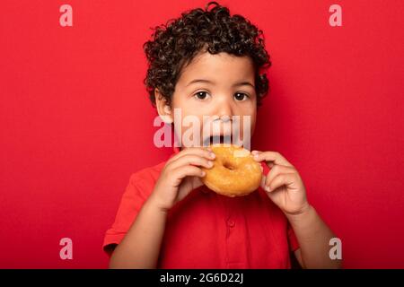 Adorable child with curly hair eating sweet tasty doughnut and looking at camera on red background Stock Photo