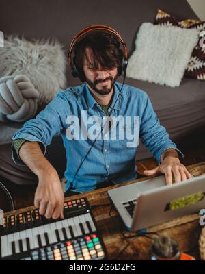 Happy young man in headphones using synthesizer and laptop at table at home Stock Photo