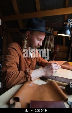 Young worker in hat sitting at his workplace and making shoes from leather in the workshop Stock Photo