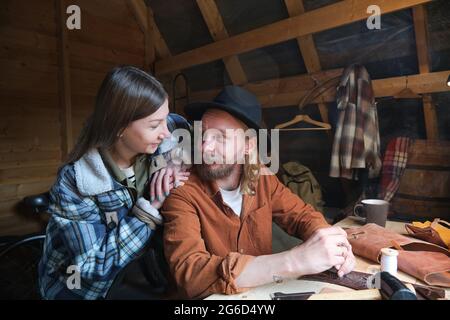 Young man sitting at the table and working with leather with young woman interrupting and talking to him in the workshop Stock Photo