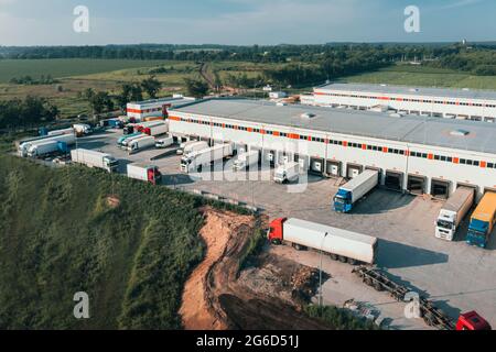 Trucks with trailers are loaded and unloaded in the cargo terminal in the morning, aerial shot. Containers and semi-trailers of trucks near the warehouse are waiting for loading goods. Stock Photo