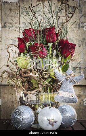 Wintry bouquet of roses in a glass vase with white and silver christmas balls and a gray woolen angel with XMAS lettering, old used wood as background Stock Photo