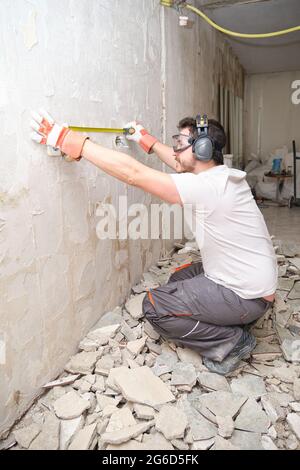 Builder man using a measuring tape at the construction site. Construction worker wearing safety gloves, glasses and earmuffs. Stock Photo