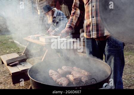 Young man frying meat on barbecue grill with woman cooking at the table in the background outdoors Stock Photo