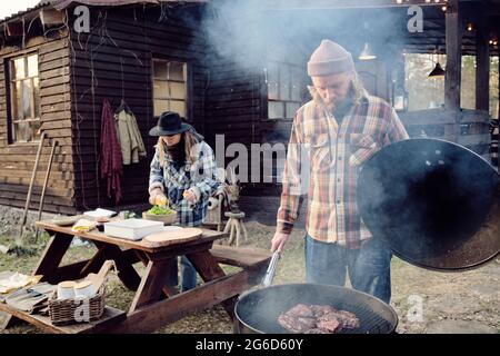 Family of two preparing dinner outdoors, man frying slices of meat on barbecue grill with woman preparing salad in the background Stock Photo