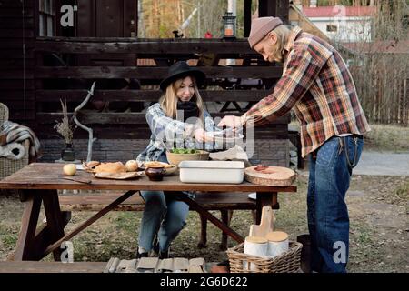 Young woman sitting at the table with man putting fried meat on the plate, they having dinner together outdoors Stock Photo