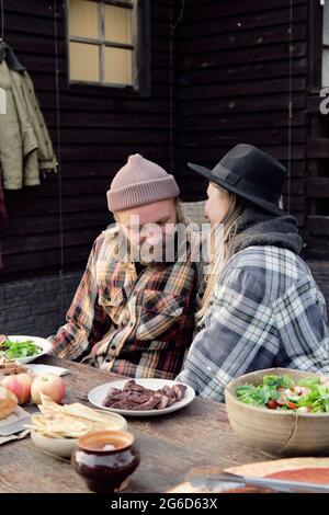 Young couple talking to each other while sitting at the table and having dinner during picnic outdoors Stock Photo