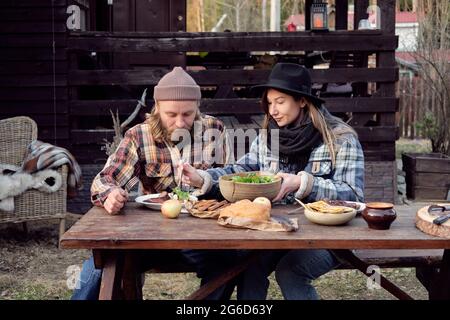 Young couple of farmers sitting at the table and eating fried meat with vegetable salad together during picnic outdoors Stock Photo