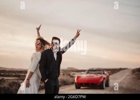 Optimistic bride and groom gesturing V sign and smiling while standing on road near red car in evening in Bardenas reales Natural Park in Navarra, Spa Stock Photo