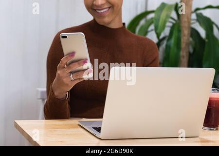 Cropped unrecognizable happy African American female freelancer browsing smartphone and smiling while sitting at table with laptop in cafe and working Stock Photo