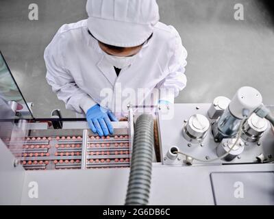 From above female factory worker in uniform operating conveyor with pills in pharmaceutical manufacturing laboratory Stock Photo
