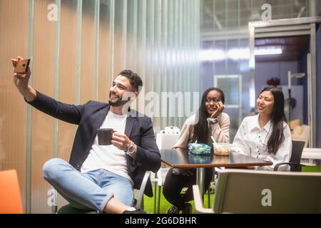Group of cheerful multiracial colleagues taking self shot on smartphone while sitting at table during lunch in workspace Stock Photo