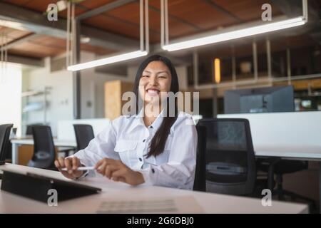 Positive Asian female employee sitting at table in workspace and looking at camera Stock Photo