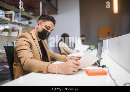 Concentrated ethnic male employee with face mask sitting at table with laptop and reading documents while working in spacious office with blurred coll Stock Photo