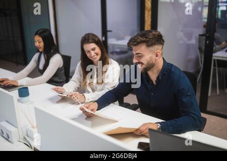 Group of cheerful multiracial coworkers sitting at table and reading documents while working together in coworking space Stock Photo