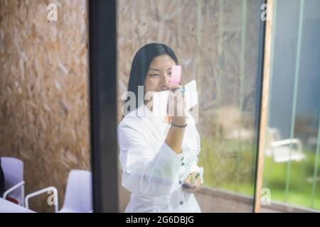 Smiling Asian female employee writing on sticky note on glass wall while working in modern office Stock Photo