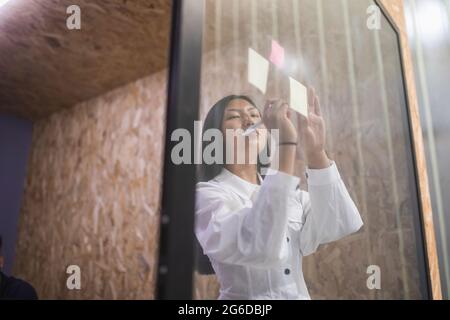 Smiling Asian female employee writing on sticky note on glass wall while working in modern office Stock Photo