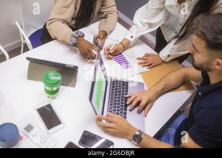 From above of group of crop colleagues gathering at table and analyzing diagrams on paper and laptop together while working on new marketing strategy Stock Photo