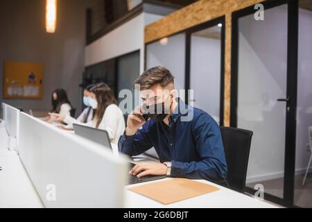 Male employee in mask typing on laptop and talking on smartphone while working in coworking space with colleagues Stock Photo