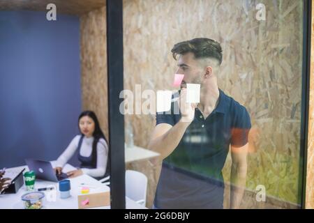 Male executive manager writing on sticky note on glass wall during brainstorm with coworkers in office Stock Photo