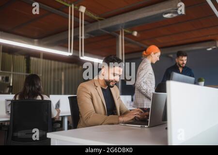 Cheerful male manager browsing netbook while sitting at table and working in coworking space with unrecognizable colleagues Stock Photo