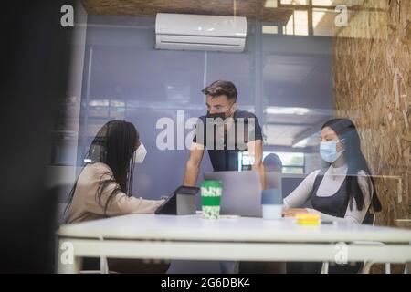 Through glass of company of multiracial coworkers in protective masks gathering at table and discussing project during business meeting Stock Photo