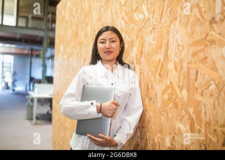 Smiling ethnic Asian female entrepreneur standing with computer near wall in coworking space while looking at camera Stock Photo