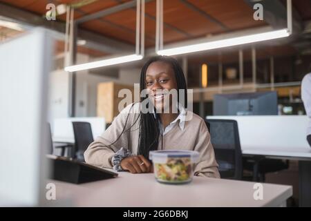 Cheerful African American female worker sitting at table with lunch box in workspace and looking at camera Stock Photo
