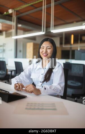 Positive Asian female employee sitting at table in workspace and looking at camera Stock Photo