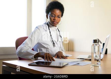Young African American female physician in white robe with stethoscope sitting at table with laptop and reading medical records while working in clini Stock Photo