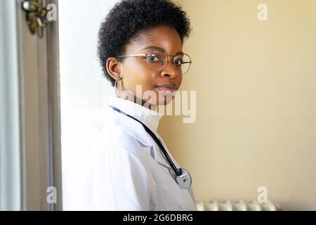 Side view of competent young African American female physician in white medical coat and eyeglasses with stethoscope looking at camera while standing Stock Photo