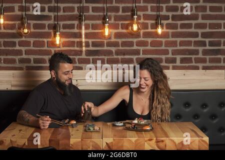 Loving couple eating appetizing sushi while sitting at wooden table in Japanese restaurant Stock Photo