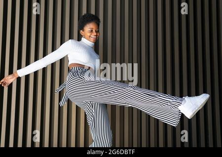 Low angle of optimistic black female in stylish clothes smiling and swinging leg while dancing against striped wall in city street Stock Photo