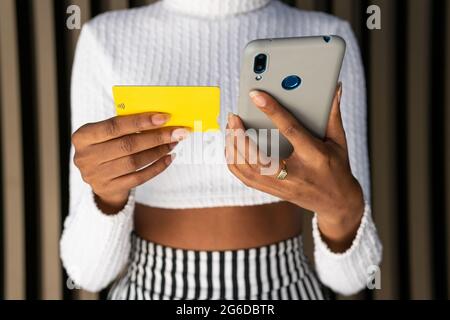 Cropped unrecognizable young African American female in trendy clothes using credit card and smartphone to pay for online order against striped wall o Stock Photo