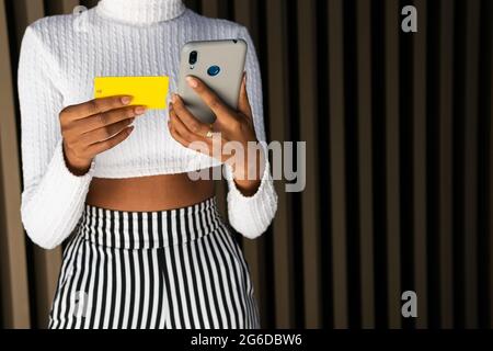Cropped unrecognizable young African American female in trendy clothes using credit card and smartphone to pay for online order against striped wall o Stock Photo